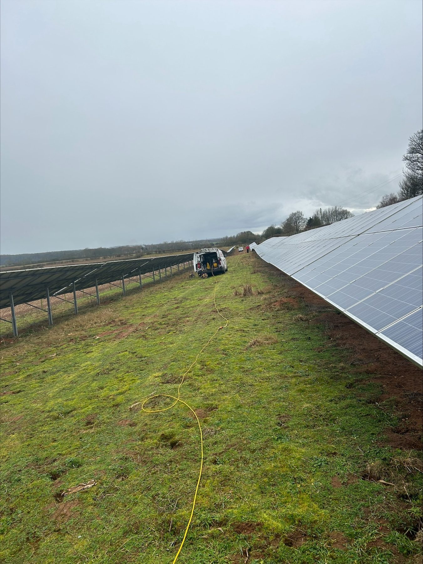 Long rows of ground-mounted solar panels on grass, with a van and hose in the distance